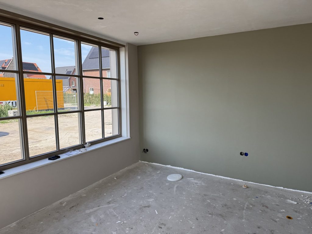 Nieuwbouwkamer met een grijsgroen geverfde muur en een breed raam dat uitkijkt op de bouwplaats